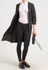Cardigan lungo nero in maglia sopra una camicia button-up rosa chiaro, abbinato a jeans skinny neri e scarpe eleganti nere lucide, mentre tiene una borsetta nera.