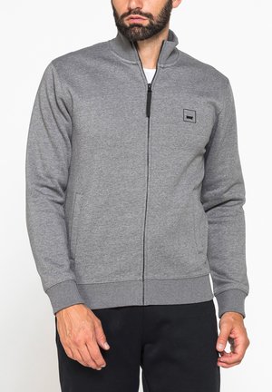 Uomo con giacca grigia con zip, polsini e orlo a coste, e pantaloni neri, in piedi davanti a uno sfondo bianco semplice.