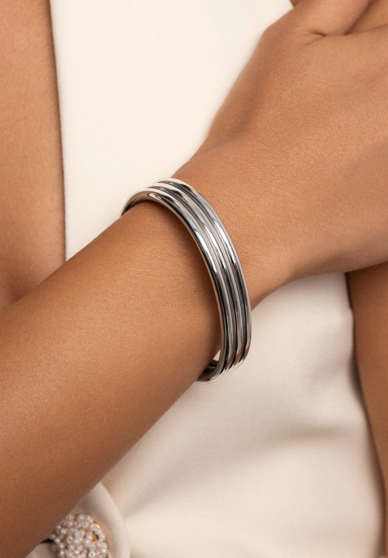 Bracciale a manica in tonalità argento con tre fasce lisce e parallele. Indossato su un polso con una tonalità di pelle chiara, presenta una finitura elegante e lucida.