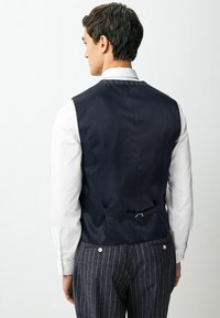 Navy blue vest with a smooth texture, featuring a back adjuster, worn over a white shirt, paired with striped trousers.