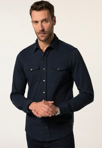 Marineblauwe blouse met lange mouwen, knopenrij, twee borstzakken, zilveren drukknoopjes en een gladde textuur. Kenmerkend zijn de puntige kraag en de getailleerde pasvorm.