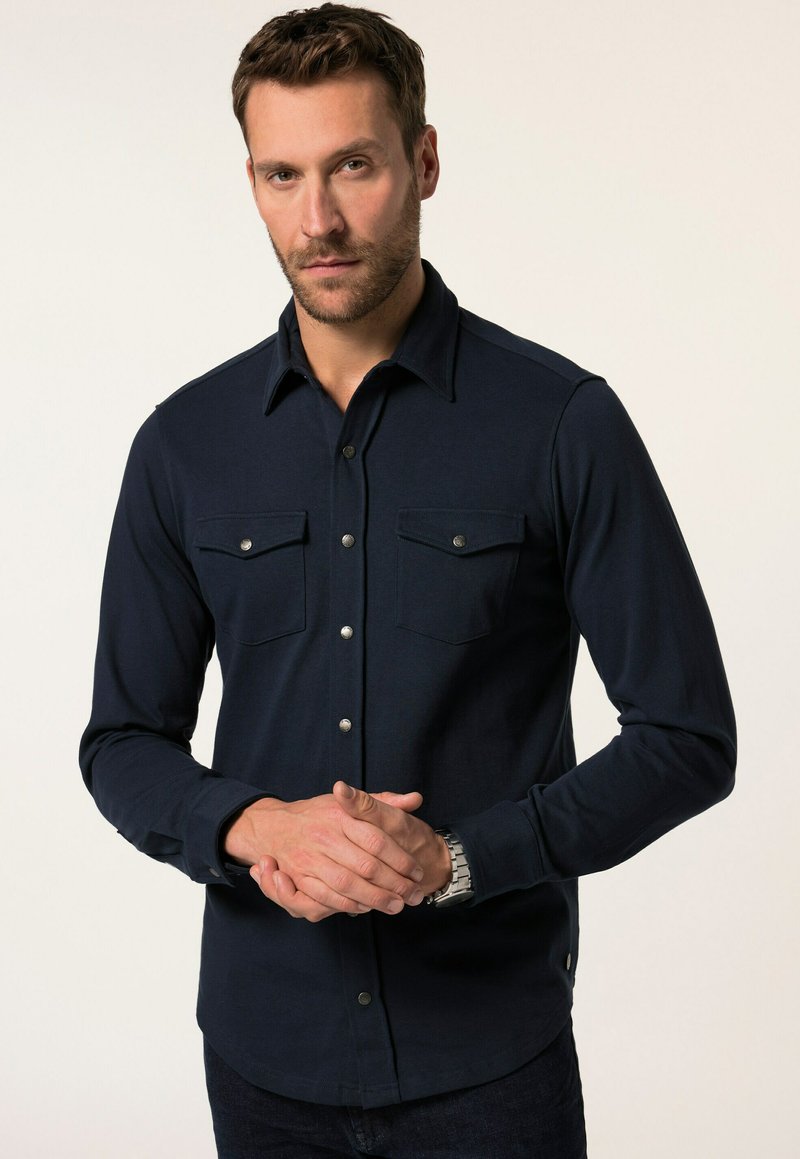 Marineblauwe blouse met lange mouwen, knopenrij, twee borstzakken, zilveren drukknoopjes en een gladde textuur. Kenmerkend zijn de puntige kraag en de getailleerde pasvorm.