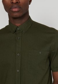Krátky rukáv, košeľa na gombíky v tmavom olivovo zelenom materiáli. Má náprsné vrecko a kontrastné gombíky, s hladkou textúrou.