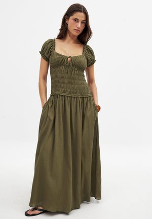 Olivgrön maxiklänning med puffärmar, smockad bodice och knytning vid halsringningen. Mjuk, texturerad tyg med sidfickor.