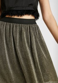 Top court noir avec des plumes, associé à une jupe mi-longue pailletée de couleur or foncé, présentant un design texturé et froncé avec une taille élastique.