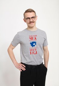 Grå bomulds t-shirt med blåt ko-grafik og røde tekster, der står "MADE WITH MILK" og "SINCE 1921." Korte ærmer, afslappet pasform, glat tekstur.