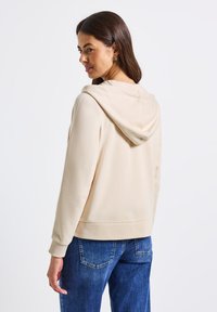 Felpa beige con cappuccio, caratterizzata da una vestibilità morbida e polsini a coste. Indossata con jeans blu, che mostrano una texture del tessuto liscia.