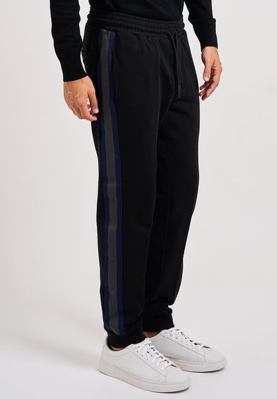 Hombre vestido con pantalones deportivos negros con rayas laterales azules y grises y zapatillas blancas, contra un fondo claro liso.