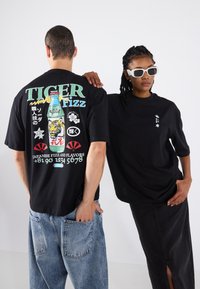 T-shirts noirs avec des imprimés graphiques : l'un présente une bouteille de soda colorée et du texte, l'autre a un petit motif blanc. Les modèles portent un jean et une jupe noire.