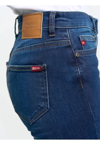 Tuvplāns uz personu, kas valkā zilas džinsa bikses ar Big Star logotipu aizmugurējā kabatā un ādas uzlīmi jostasvietā.