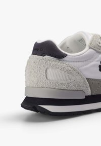 Zapatilla blanca con acentos de gamuza y malla gris, collar acolchado y suela negra. Presenta un bordado del logo en el talón y detalles texturizados.