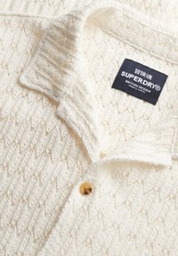 Camisa de punto blanca texturizada con un patrón de espiga, un cuello y un botón dorado. Muestra una etiqueta de marca negra que dice "Superdry."