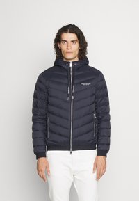 Marineblauer Pufferjacke mit Kapuze, verfügt über einen Reißverschluss vorne, elastische Bündchen und zwei Seitentaschen. Glattes Gewebe mit horizontalem Steppmuster.