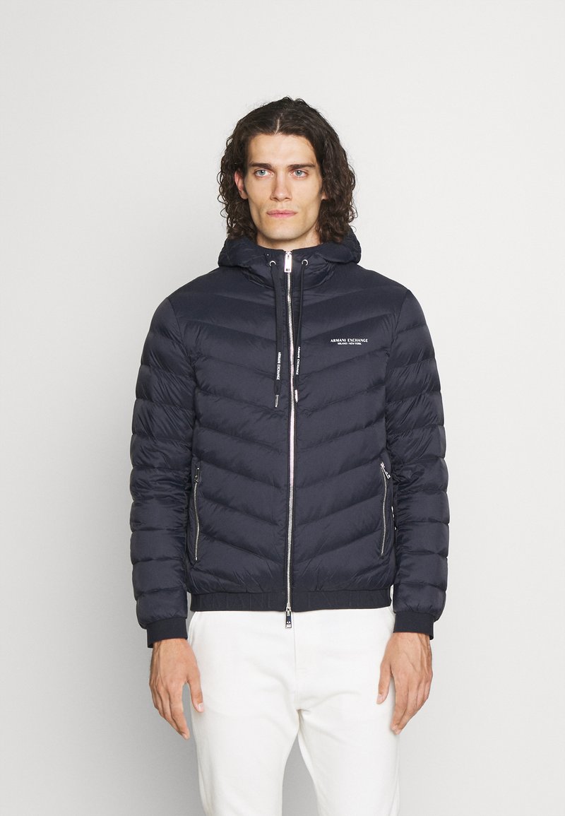 Marineblauer Pufferjacke mit Kapuze, verfügt über einen Reißverschluss vorne, elastische Bündchen und zwei Seitentaschen. Glattes Gewebe mit horizontalem Steppmuster.
