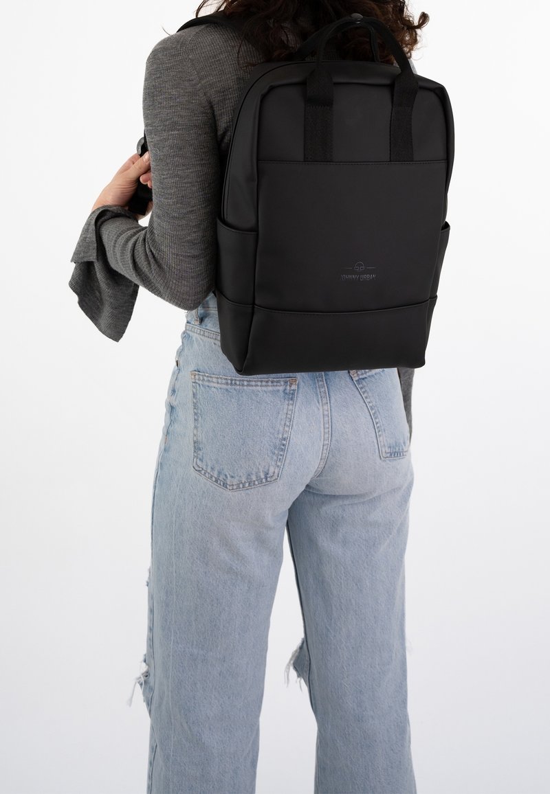 Personne portant un jean bleu clair et un haut gris à manches longues avec des poignets évasés, portant un sac à dos rectangulaire noir dans son dos.