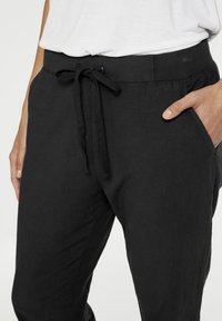 Kuvion vyötäröstä puoliväliin reisiä, jossa henkilö pukeutuu mustiin naruhousuihin ja valkoiseen paitaan, oikea käsi osittain housujen taskussa.