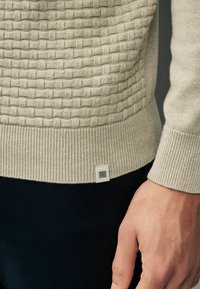 Pull en maille beige avec motif tressé texturé sur le torse, poignets et ourlet côtelés, petite étiquette logo en tissu, porté avec un pantalon foncé.