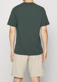 Tmavě zelené bavlněné tričko s kulatým výstřihem, krátkými rukávy a volným střihem, v kombinaci se světlými béžovými šortkami. Žádné viditelné vzory ani akcenty.