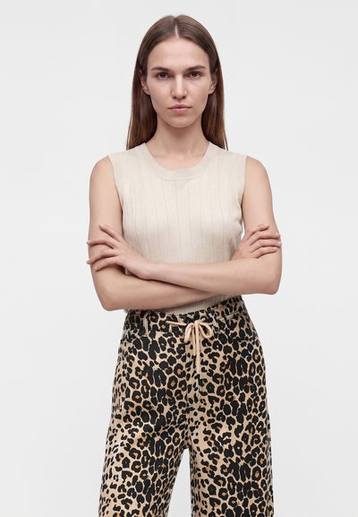 Femme aux longs cheveux bruns et expression neutre, portant un top beige sans manches et un pantalon imprimé léopard avec cordon, bras croisés.