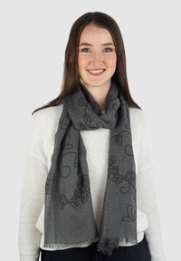 Grijze sjaal met zwarte bloemenpatronen. De stof lijkt licht en textuur te hebben, met franjes aan de randen. Losjes om de nek gedragen.
