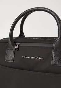 Zwarte stoffen tas met gestructureerde leren handvatten, een ritsvak en het "TOMMY HILFIGER" logo in het wit. Schoon, minimalistisch ontwerp.