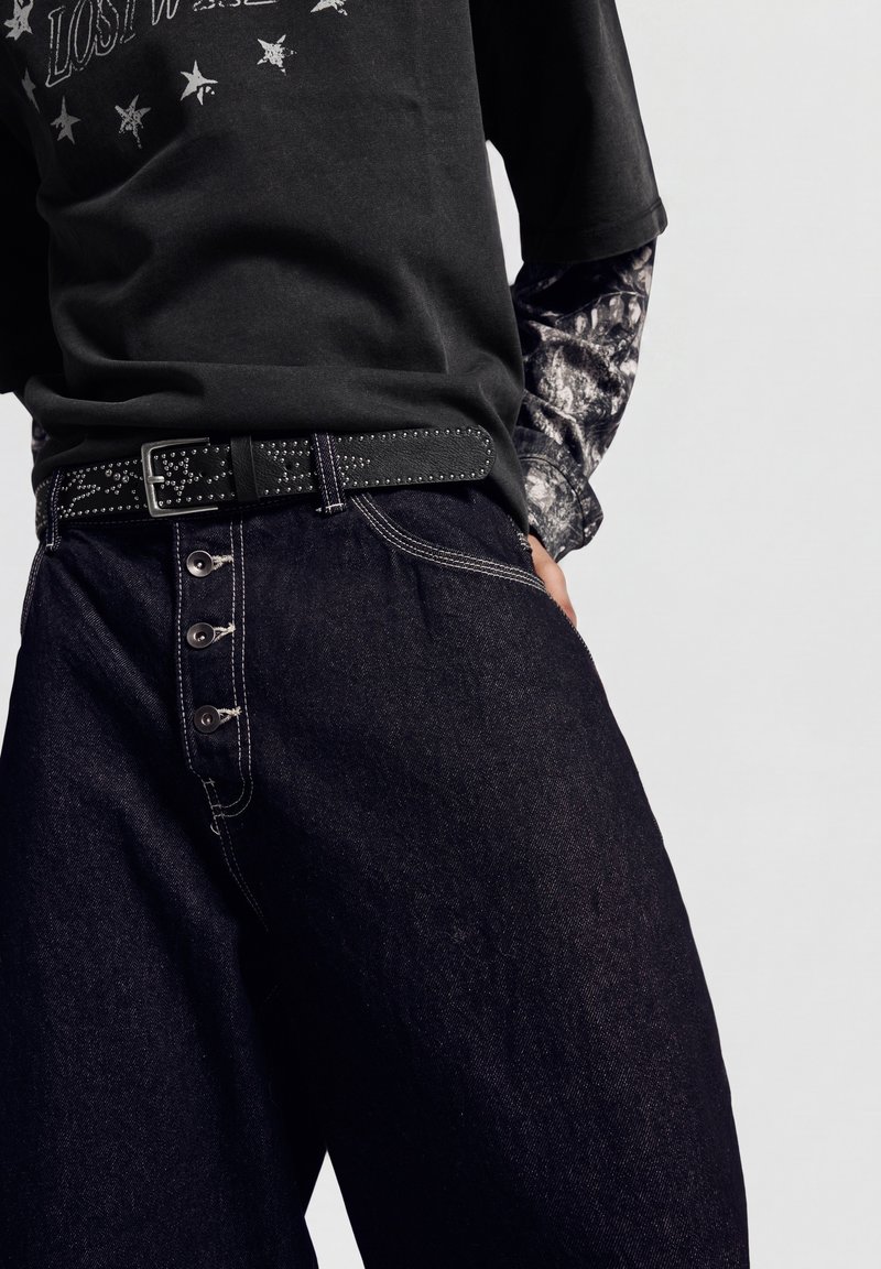 Personne portant une ceinture noire cloutée, un t-shirt noir à motif graphique sur une chemise à manches longues avec un imprimé tête de mort, et un jean taille haute foncé avec des boutons visibles.