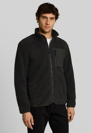Mann trägt schwarze Fleecejacke mit Reißverschluss, mit einer Reißverschlusstasche auf der Brust und Seitentaschen, steht vor einem schlichten hellen Hintergrund.