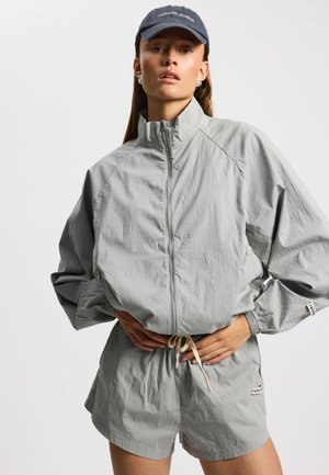 Joven mujer con chaqueta con cierre de color gris claro y shorts a juego, gorra azul marino con texto blanco, de pie con las manos en la cintura sobre un fondo blanco.
