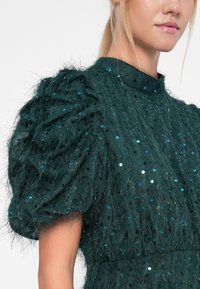 Dark teal dress with textured, fluffy sleeves, adorned with sequins. Features a high neckline and gathered detailing.