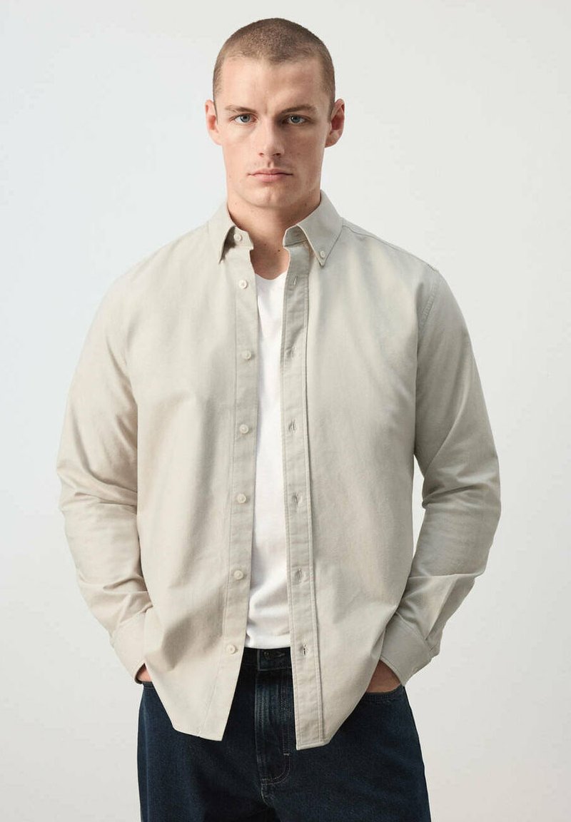 Jeune homme aux cheveux courts portant une chemise beige boutonnée sur un t-shirt blanc, les mains dans les poches d'un jean foncé, fond neutre.