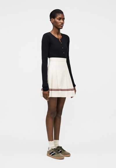 Γυναίκα που φοράει μαύρο πουκάμισο με κουμπιά και μακριά μανίκια, λευκή πλισέ φούστα με κόκκινες ρίγες, λευκές κάλτσες και ελιά πράσινα παπούτσια, στεκόμενη στο πλάι.