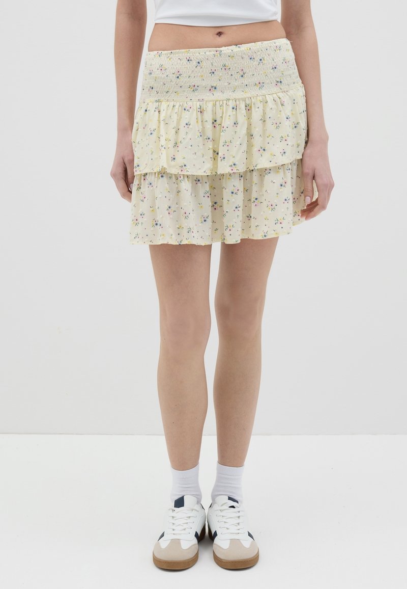 Jupe tiered jaune à fleurs avec une taille smockée, ornée de petites fleurs multicolores et d'une texture légère et transparente. Portée avec des baskets blanches.