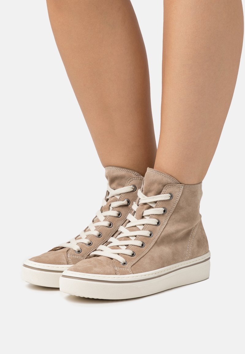 Jambes portant des baskets montantes en daim beige avec des lacets blancs et des semelles épaisses en caoutchouc blanc sur un fond neutre.
