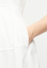 Bras à la peau claire avec des taches de rousseur reposant dans la poche latérale d'un vêtement blanc.