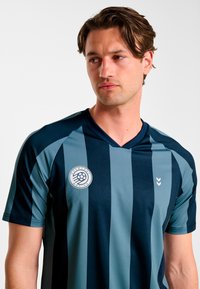 Granatowo-turkusowa sportowa koszulka w paski z dekoltem w serek, teksturowana tkanina oraz okrągłe logo na piersi z kontrastującymi akcentami kolorystycznymi.