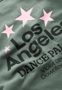Tejido verde con gran texto negro que dice "Los Ángeles" y "PALACIO DEL BAILE". Dos estrellas rosas están encima del texto, con detalles bordados.