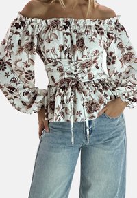 Kobieta w białej bluzce z odkrytymi ramionami z brązowym kwiatowym wzorem i wiązanym gorsetem w talii, zestawiona z jasnoniebieskimi, wysokim stanem dżinsami.