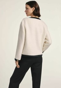 Biała teksturowana kurtka z czarnym kołnierzykiem i mankietami, charakteryzująca się luźnym krojem i złotymi guzikami na rękawach. Noszona na czarnych spodniach.