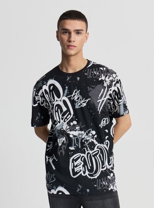 Giovane uomo con capelli corti che indossa una maglietta nera oversize con testo in stile graffiti bianco e grigio e motivi di schizzi di vernice, in piedi su uno sfondo grigio.