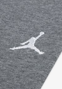 Grijze stof met een gladde textuur, voorzien van een witte geborduurde Jumpman-logo. Het ontwerp benadrukt sportieve branding.