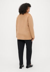 Καφέ σακάκ�ι από μαλλί με χαλαρή εφαρμογή, με στρογγυλή λαιμόκοψη και μακριά μανίκια, φορεμένο πάνω από μαύρα παντελόνια και μαύρες ψηλές γόβες.