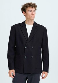 Junger Mann mit lockigem Haar, der ein schwarzes doppelt geknöpftes Blazer über einem weißen Hemd trägt und zur Seite blickt, vor einem einfarbigen Hintergrund.