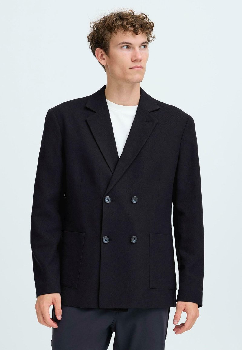 Junger Mann mit lockigem Haar, der ein schwarzes doppelt geknöpftes Blazer über einem weißen Hemd trägt und zur Seite blickt, vor einem einfarbigen Hintergrund.