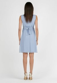 Helles blaues Kleid mit tailliertem Schnitt und Schleifendetail auf der Rückseite. Glatter, fließender Stoff; knielang. Das Model trägt goldene High-Heels.