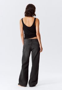 Donna con capelli scuri alla spalla, indossa una canottiera nera e jeans larghi neri, in piedi di spalle su uno sfondo bianco uniforme.