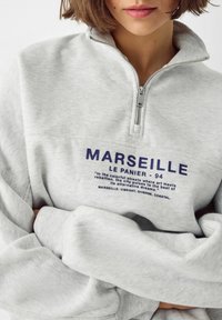 Graues Fleece-Sweatshirt mit einem halben Reißverschluss am Kragen, das einen blauen Grafikdruck mit Text über Marseille und einen gerippten Saum zeigt.