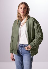 Oliivinvihreä bomber-takki, jossa on sileä pinta, resorit hihansuissa, sivutaskut ja vetoketjusuljenta, yhdistettynä valkoiseen paitaan ja sinisiin farkkuihin.