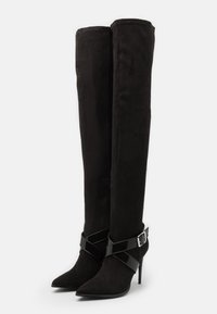 Botas negras de ante por encima de la rodilla, con punta afilada y tacón de aguja. Equipadas con una correa de charol brillante en forma de cruz.