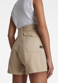 Kaki kraťasy s vysokým pasem, s kapesními zadními kapsami, kontrastním prošitím a malou značkovou cedulkou na pravé kapse.