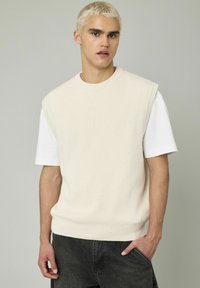 Pull en tricot crème sans manches sur une chemise blanche, avec un col rond et un ourlet côtelé, associé à un jeans gris foncé.